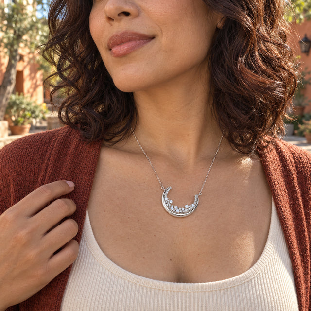 A woman with dark curls wears the silver crescent necklace above a cream top and rust cardigan in a sunny courtyard.