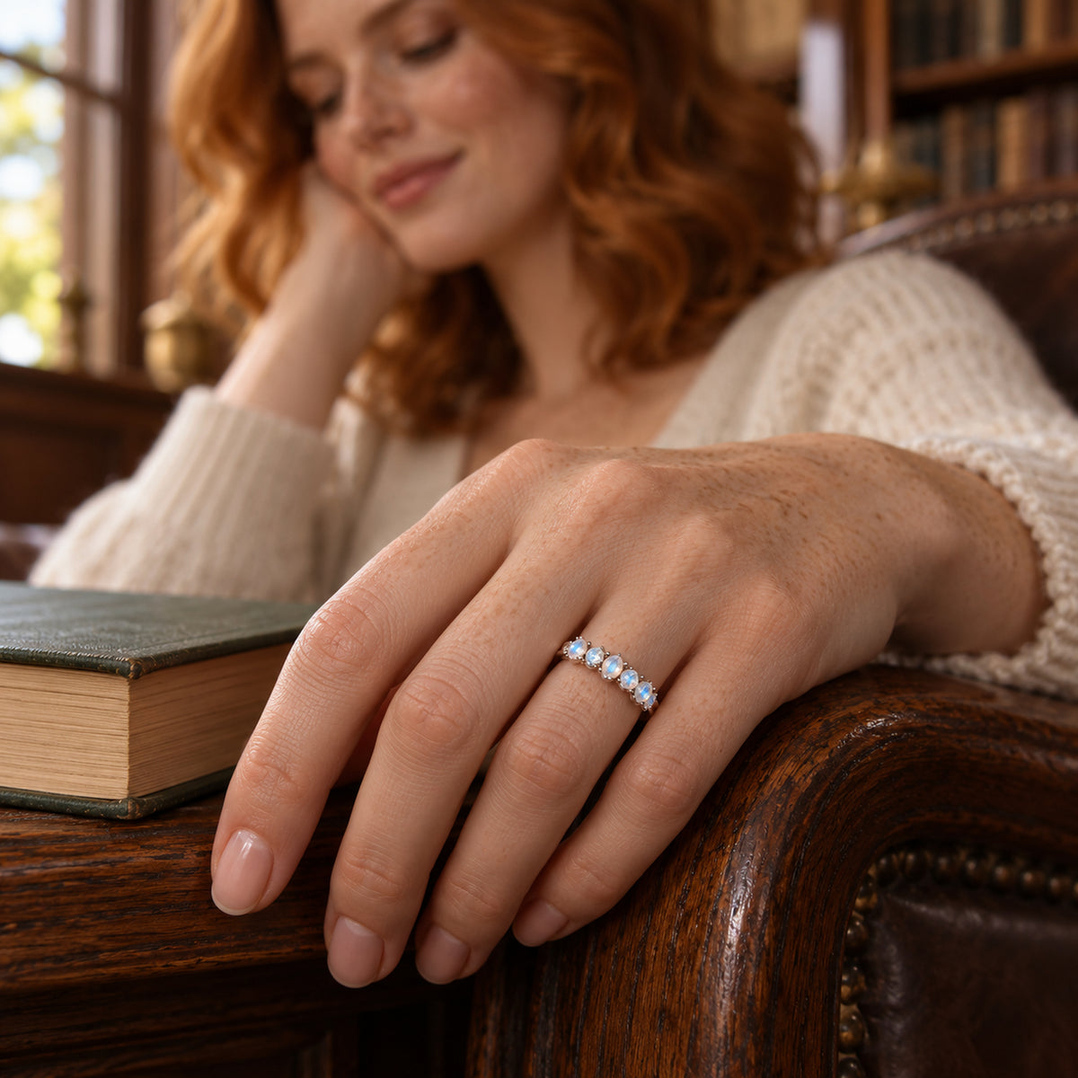 A white gold moonstone and diamond ring is shown on a freckled hand beside a book and a wooden chair arm.