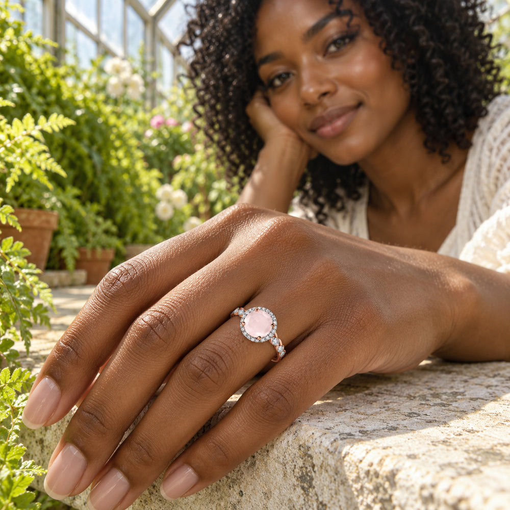 A rose gold ring with a pale pink stone and diamond halo is worn on a hand resting on a stone bench in a greenhouse.