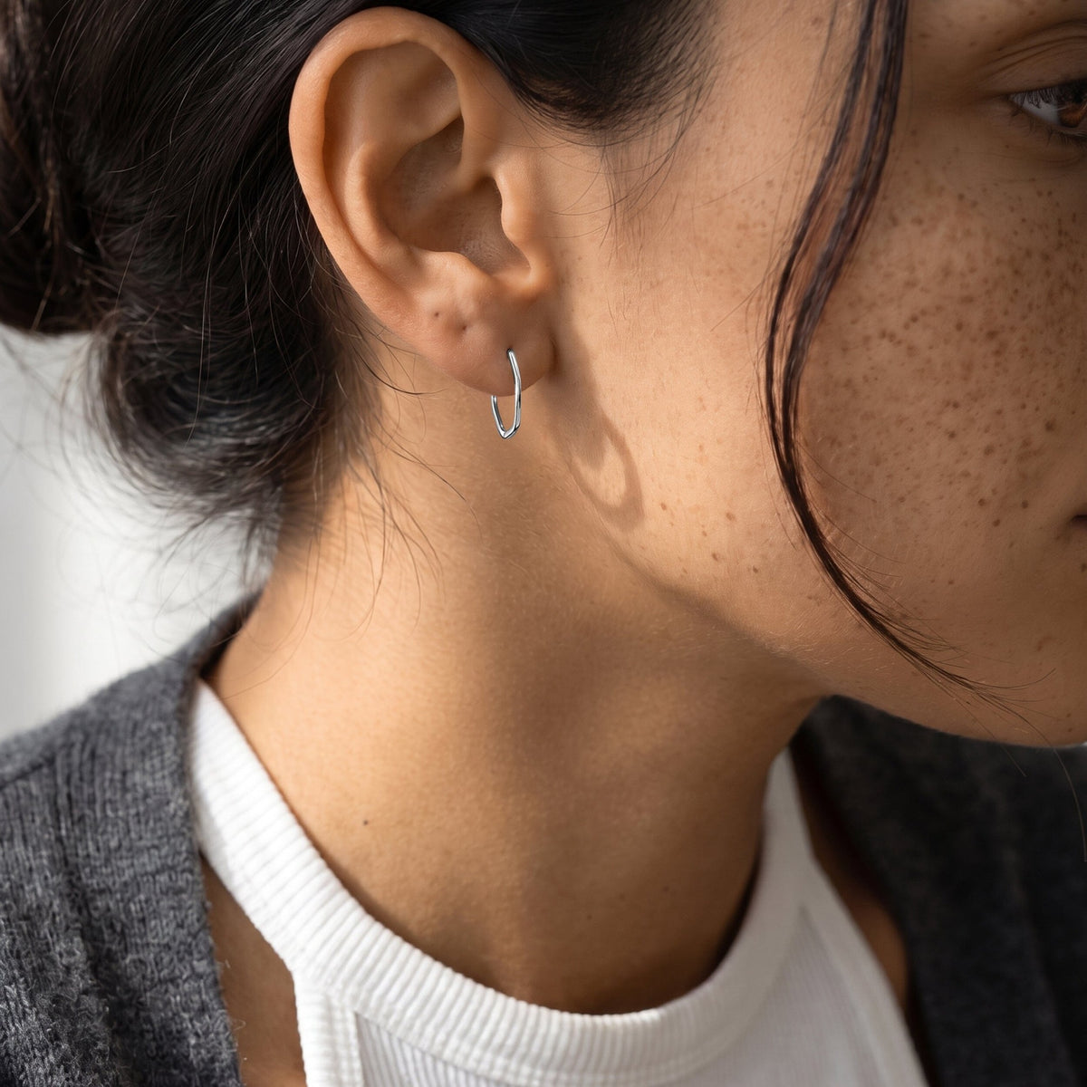 Close-up of a person wearing a small wavy hoop earring beside dark hair and a freckled cheek.