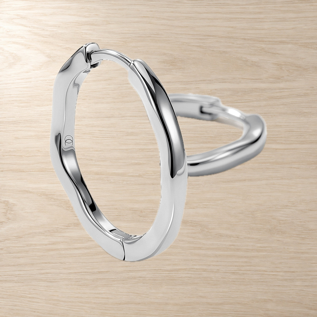 Close-up product view shows two polished irregular oval hoops overlapping on a pale wood surface.