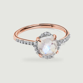 A rose gold ring with a cushion-shaped moonstone and diamond halo rests on a light gray surface.
