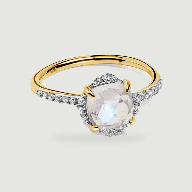 A yellow gold ring is centered on pale gray with a cushion-cut moonstone and a narrow diamond-set band.