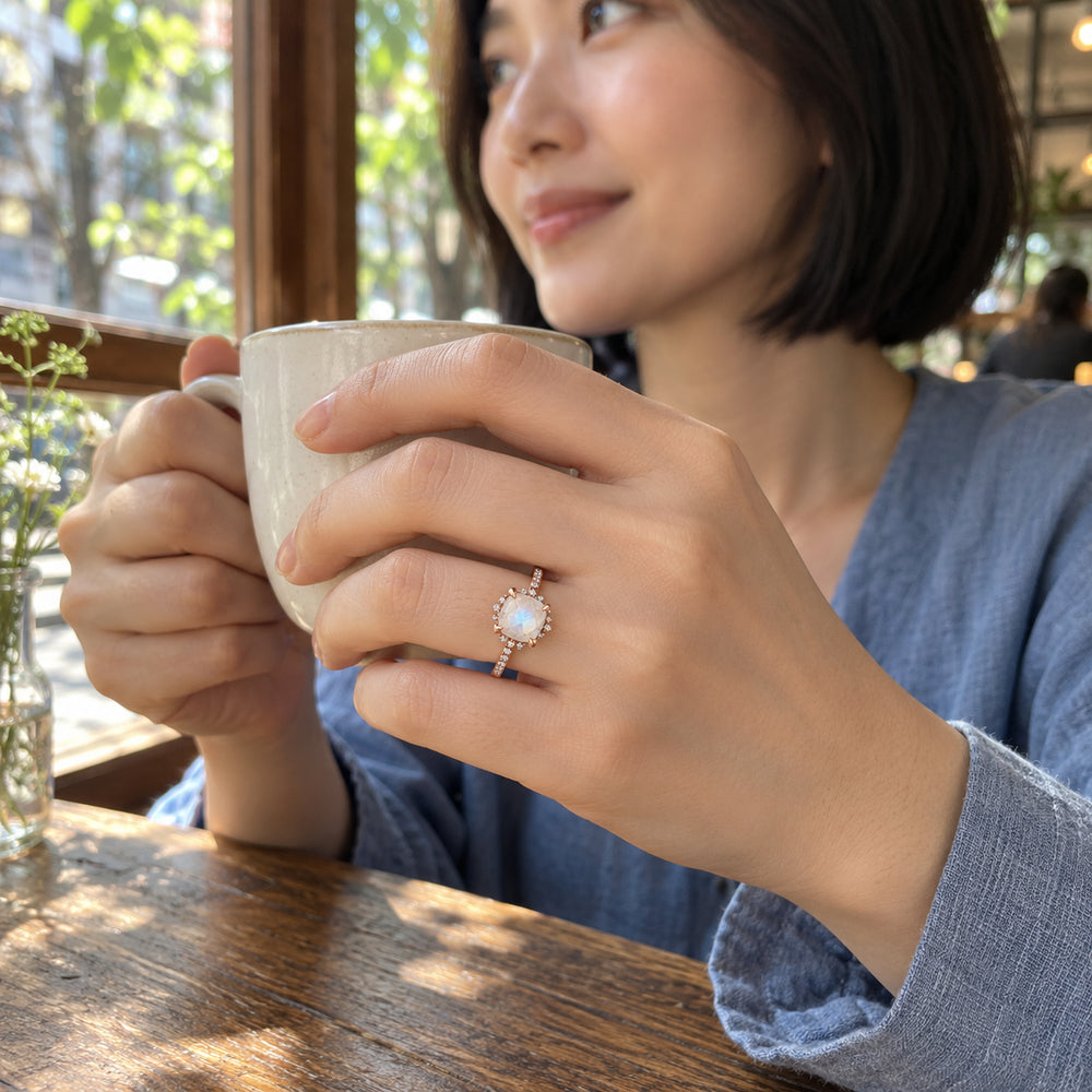 A rose gold moonstone ring is worn on a hand holding a ceramic cup beside a cafe window.
