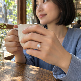 A rose gold moonstone ring is worn on a hand holding a ceramic cup beside a cafe window.