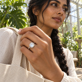 A white gold moonstone ring catches the sunlight on a hand holding a woven tote in a leafy conservatory.