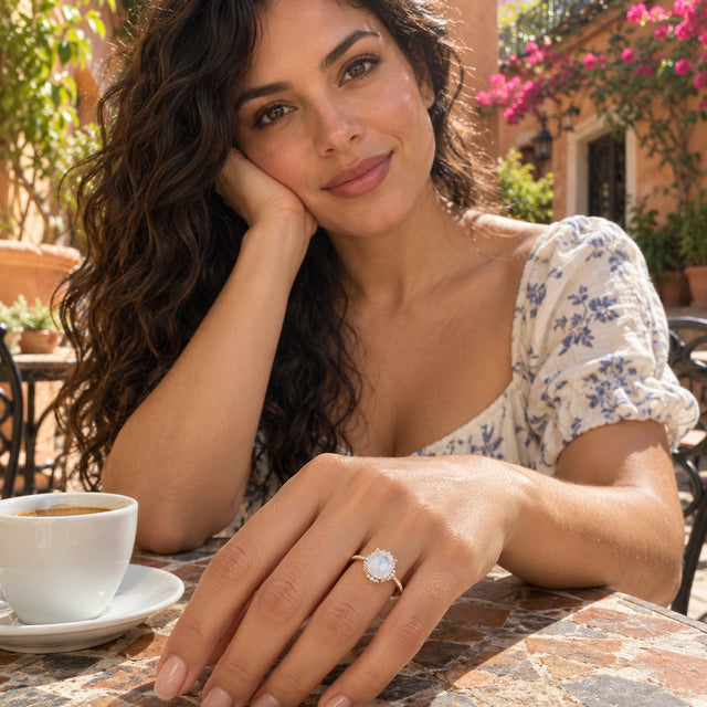 A yellow gold moonstone ring is shown on a hand resting beside a coffee cup on an outdoor bistro table.