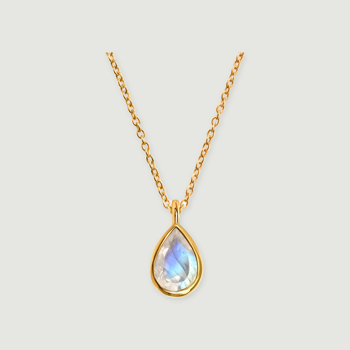 A yellow gold necklace shows a blue-flashing pear-shaped moonstone in a smooth bezel on a pale gray surface.