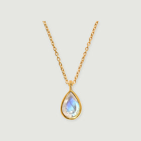 A yellow gold necklace shows a blue-flashing pear-shaped moonstone in a smooth bezel on a pale gray surface.