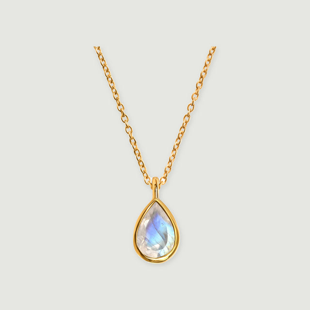 A yellow gold necklace shows a blue-flashing pear-shaped moonstone in a smooth bezel on a pale gray surface.