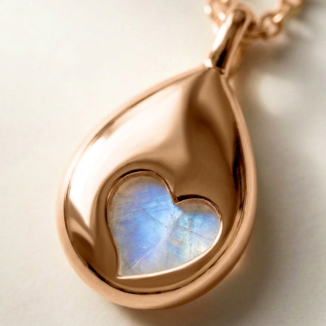 A rose gold pendant lies on a pale stone tabletop with its heart-shaped reverse opening and blue stone detail in view.