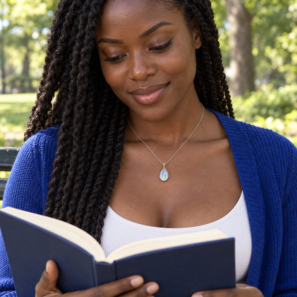 A woman in a blue cardigan wears a silver pear-shaped moonstone necklace while reading on a park bench.
