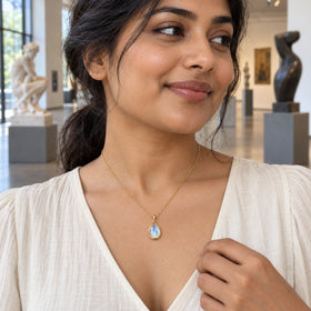 A yellow gold moonstone pendant rests above a cream blouse as a woman stands in a sunlit sculpture gallery.