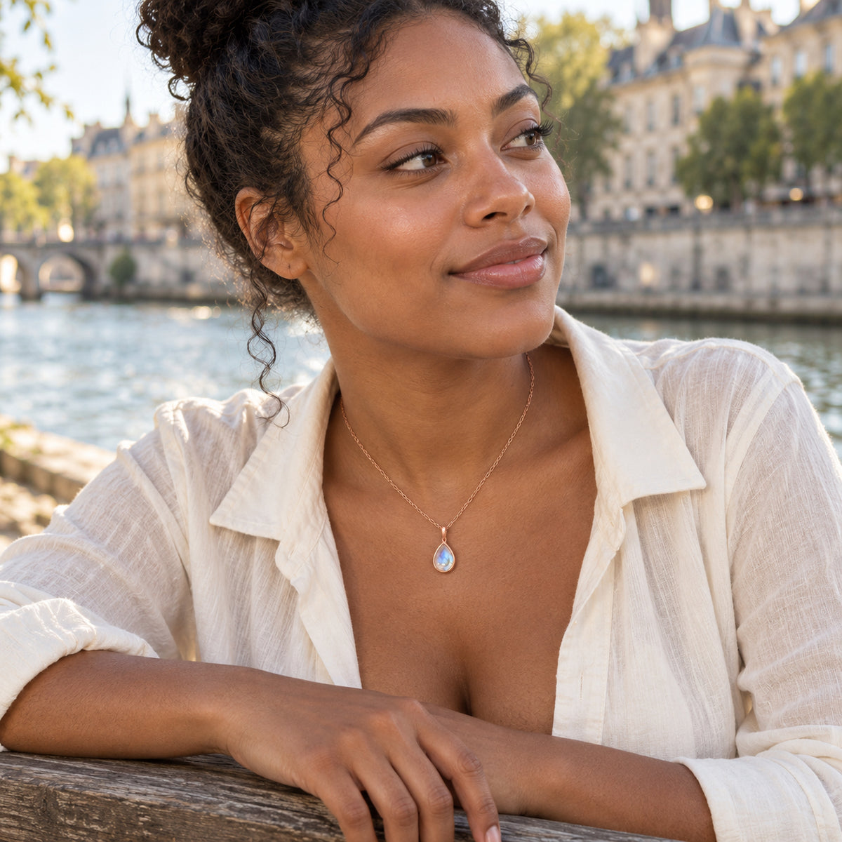 A woman with curly hair wears a rose gold moonstone necklace and an ivory shirt beside a river.
