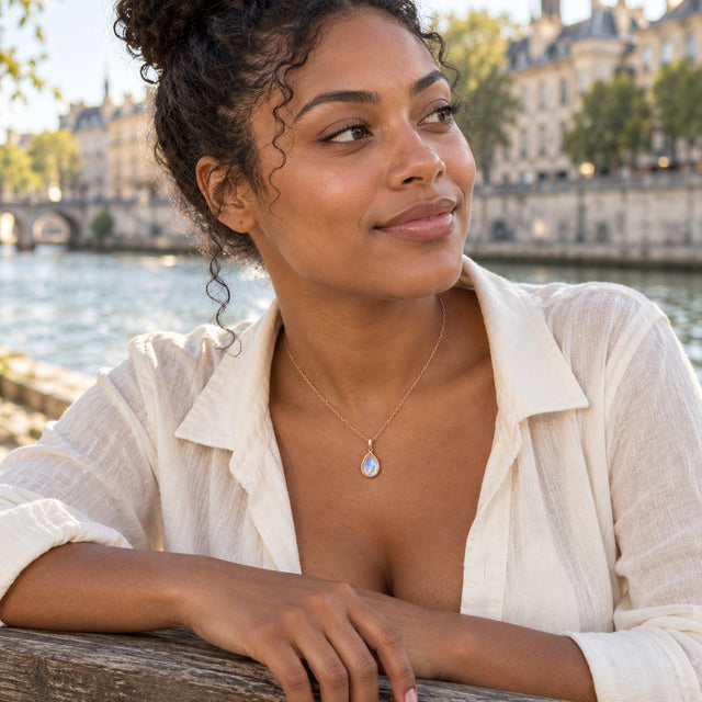 A woman with curly hair wears a rose gold moonstone necklace and an ivory shirt beside a river.