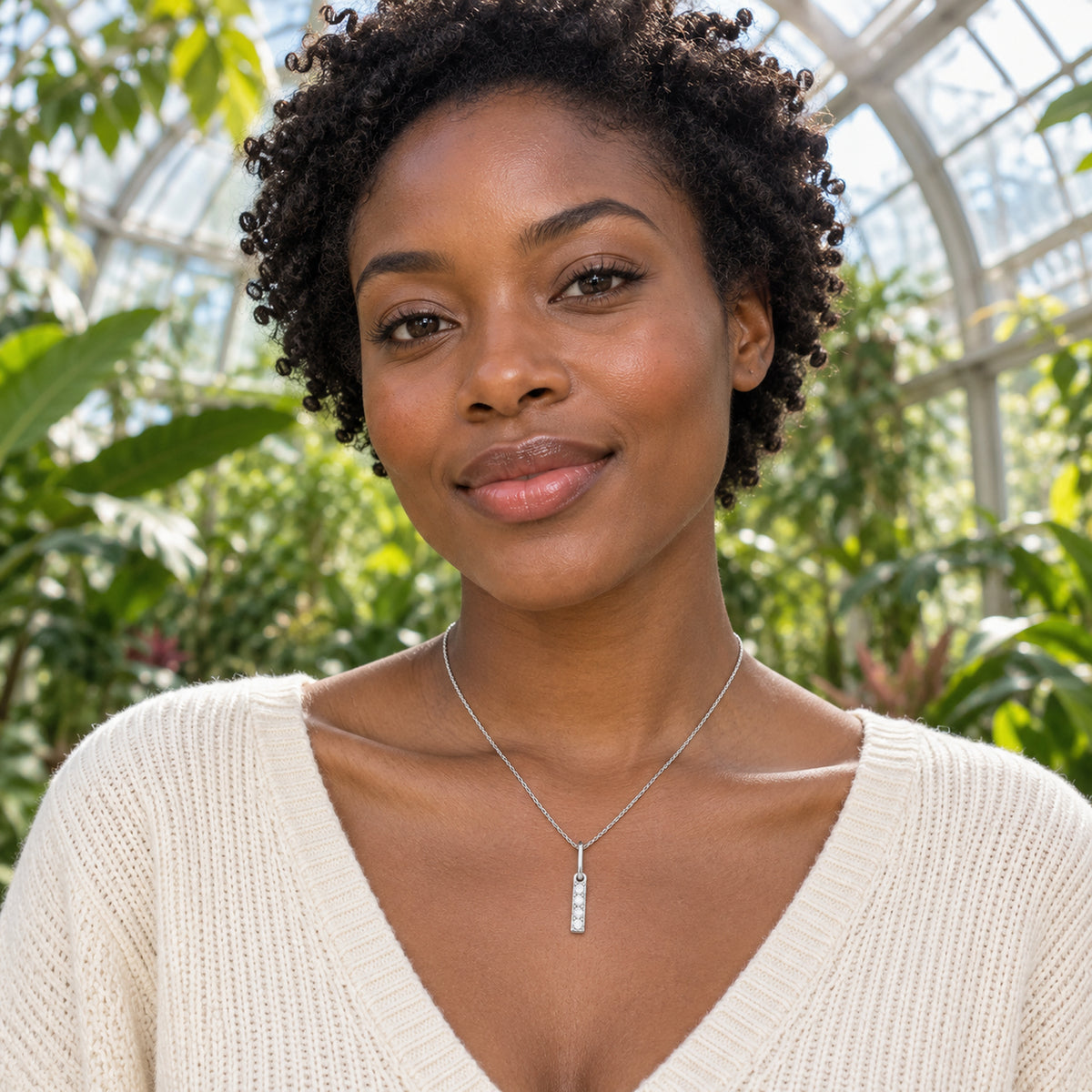 In a glass conservatory, a woman in a cream knit top wears a narrow silver pendant set with pale round stones.
