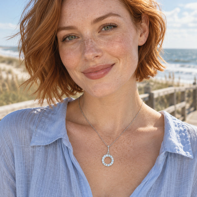 A circular silver pendant with pale stones hangs above a blue shirt against a blurred seaside boardwalk.