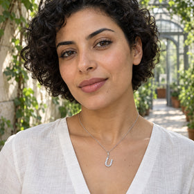 A woman with short dark curls wears a silver U pendant above an ivory linen blouse among greenhouse foliage.