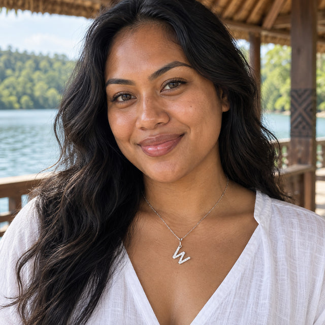 A silver zigzag pendant with pale stones hangs above a white linen blouse at a lakeside wooden pavilion.