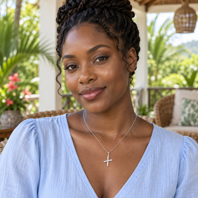A woman in a blue V-neck blouse wears a silver pendant with intersecting stone-set arms on a tropical porch.