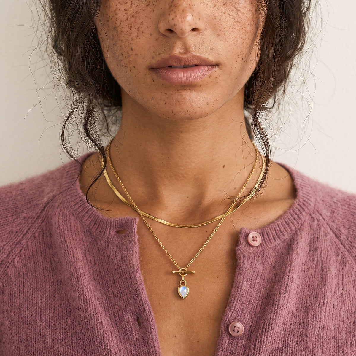 Close-up of a person in a mauve cardigan wearing the pear-shaped pendant beneath layered chains.