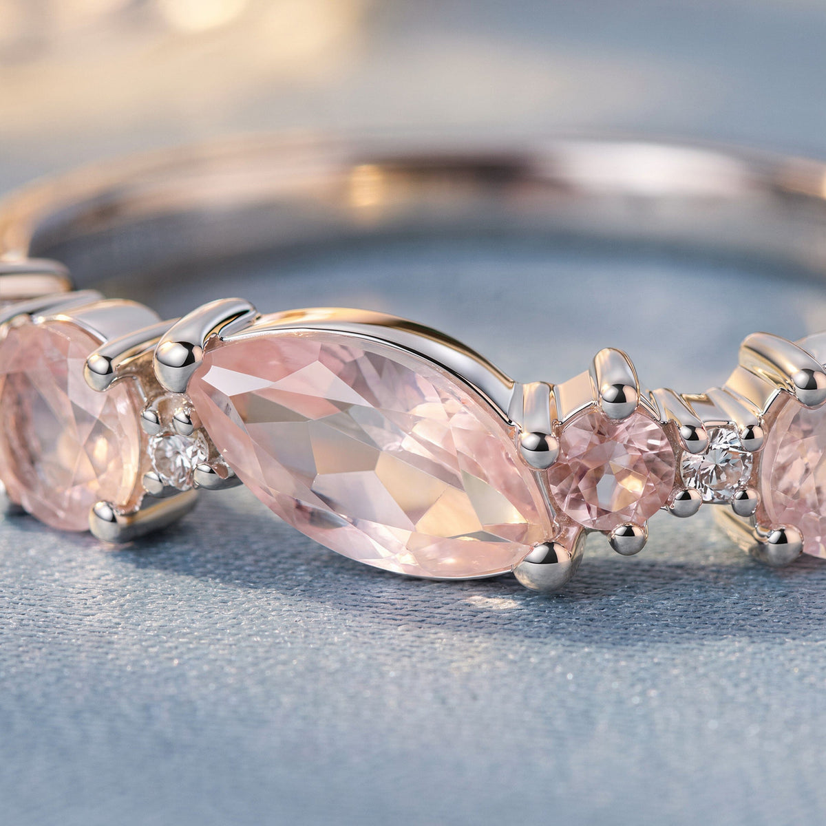 A faceted pink marquise stone and small clear accents sit in polished silver settings on a textured tabletop.
