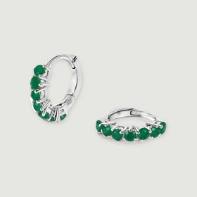 A pair of white rhodium plated hoops shows green gemstones and polished hinged closures on light gray.