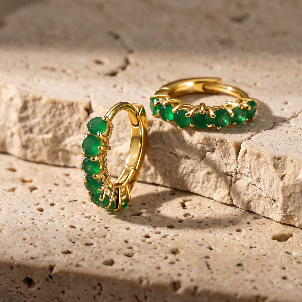 Yellow gold plated green onyx hoops sit upright and flat on textured beige stone.