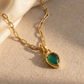 A yellow gold plated green pendant and elongated chain links rest across beige linen beside folded ivory fabric.