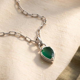 A green pear pendant on a white rhodium plated chain lies on textured ivory cloth beside an olive fabric edge.