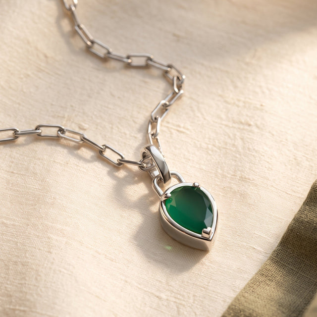 A green pear pendant on a white rhodium plated chain lies on textured ivory cloth beside an olive fabric edge.