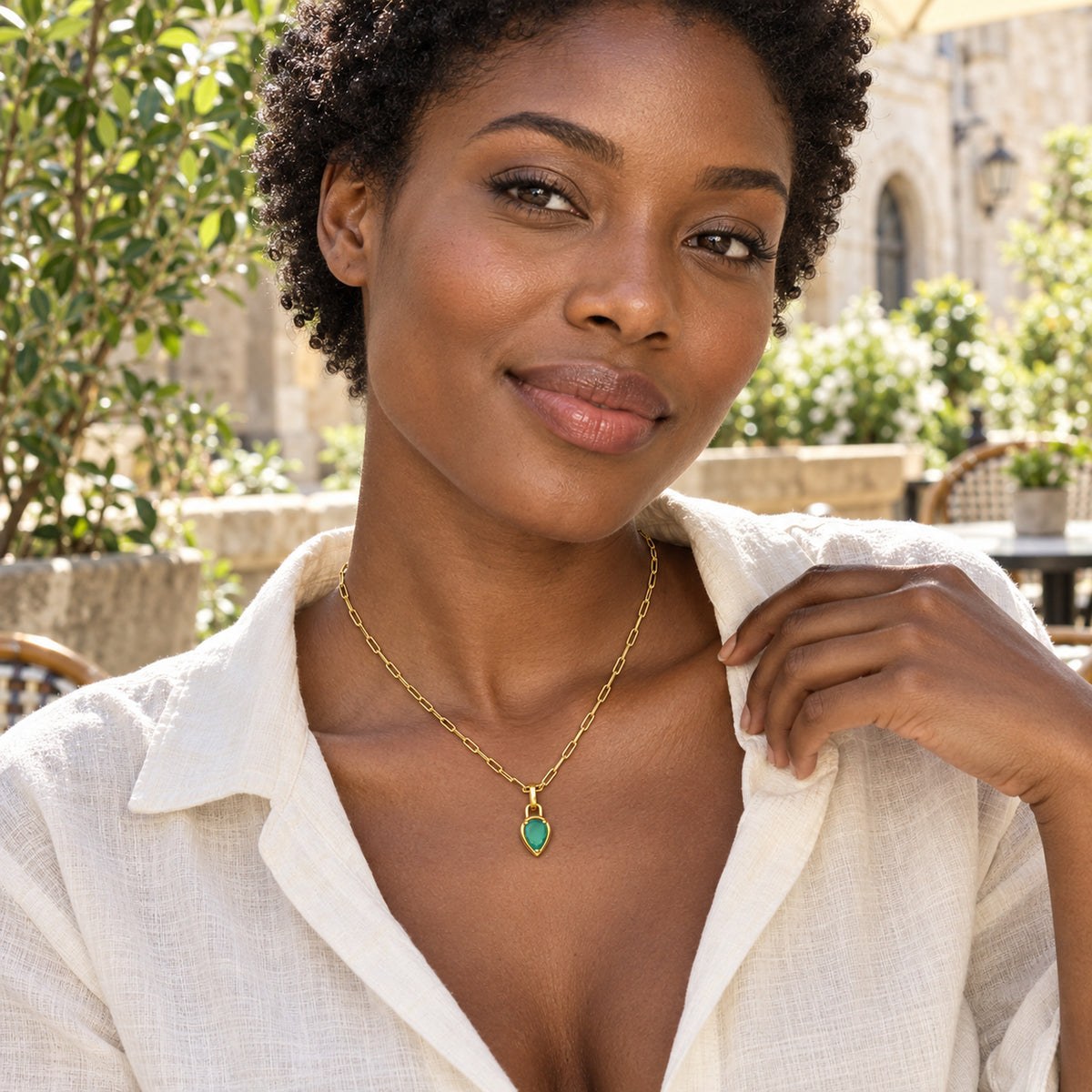 A woman with short coiled hair wears the yellow gold plated green pendant necklace with a cream linen blouse on a sunlit cafe terrace.