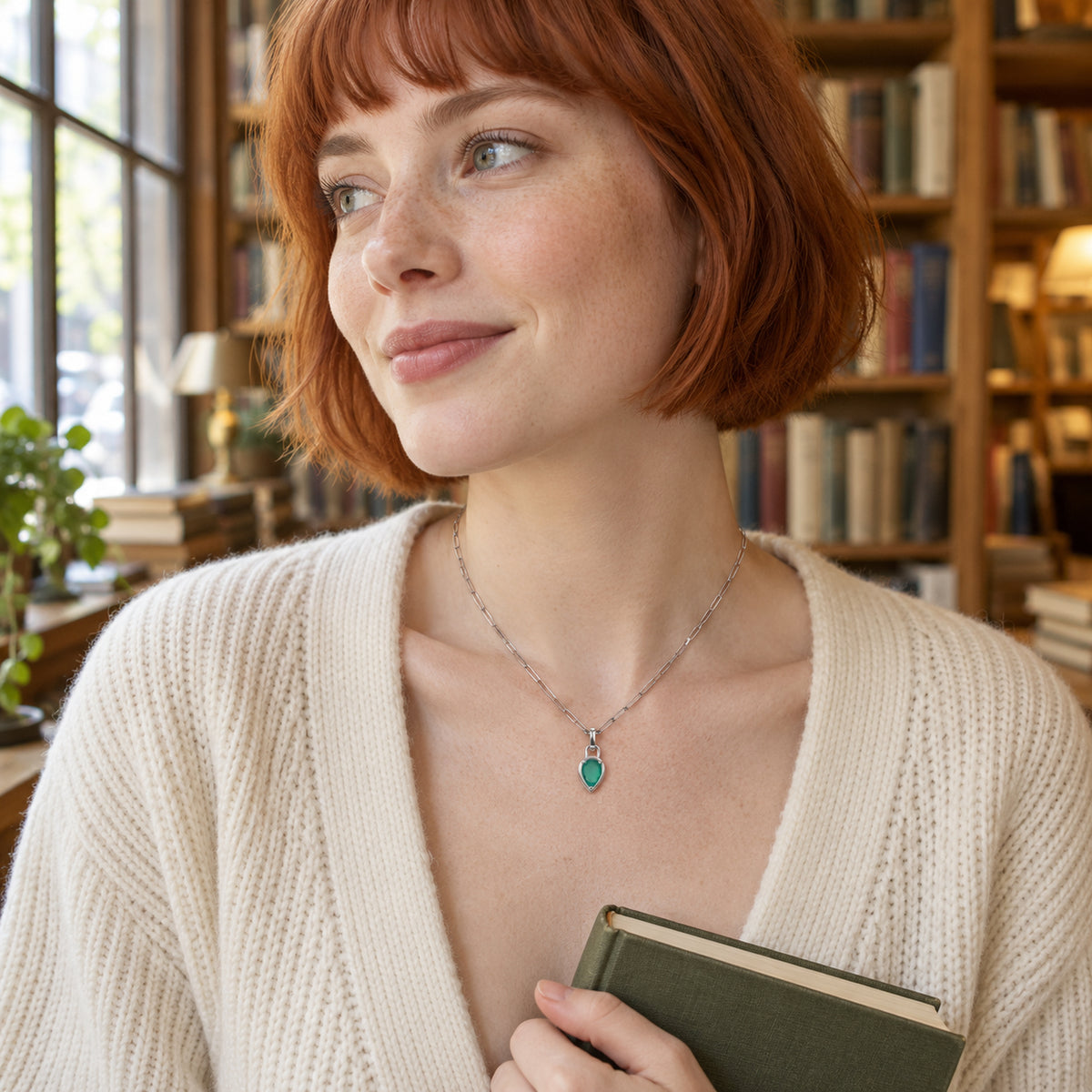 A woman with a copper-red bob wears the white rhodium plated green pendant necklace and an ivory cardigan while holding a book beside a window.
