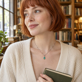 A woman with a copper-red bob wears the white rhodium plated green pendant necklace and an ivory cardigan while holding a book beside a window.