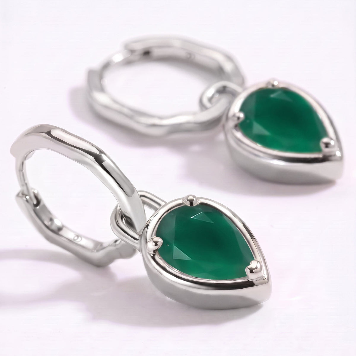 The silver-colored hoops and pear-shaped green stones rest on a clean pale surface in close-up.