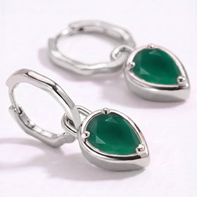 The silver-colored hoops and pear-shaped green stones rest on a clean pale surface in close-up.