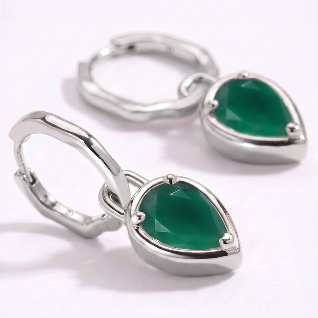 The silver-colored hoops and pear-shaped green stones rest on a clean pale surface in close-up.