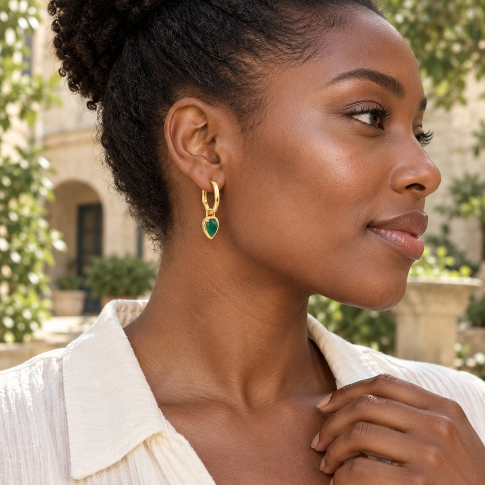 A woman with coily hair wears a gold plated green pear-drop hoop in a leafy courtyard.