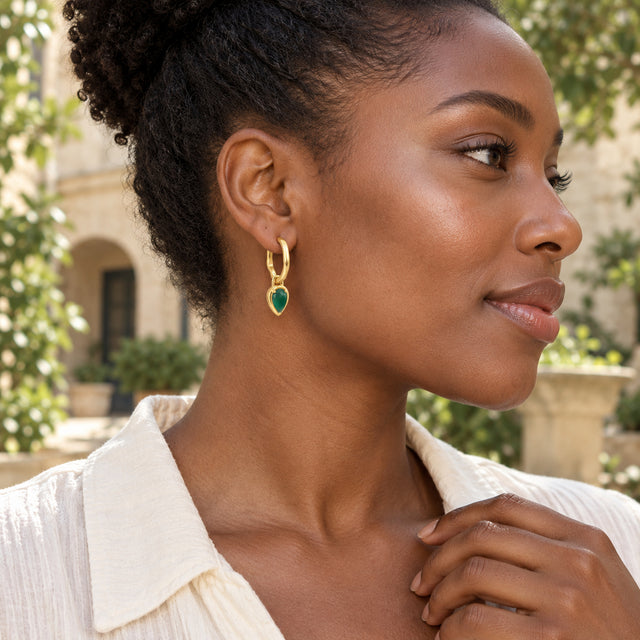 A woman with coily hair wears a gold plated green pear-drop hoop in a leafy courtyard.