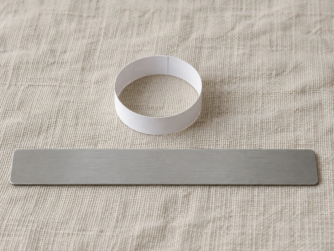 Natural window light illuminates a plain paper ring-sizing band beside an unmarked ruler showing its width.