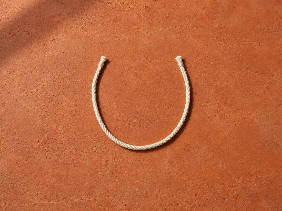 Natural window light illuminates one loose silk cord forming an open U on an abstract linen board, no body silhouette.