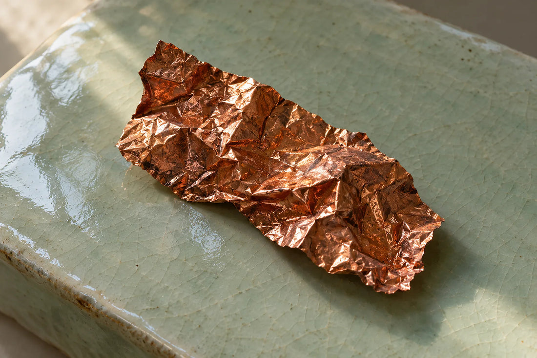 Natural window light illuminates one crumpled copper foil fragment with red metallic sheen.