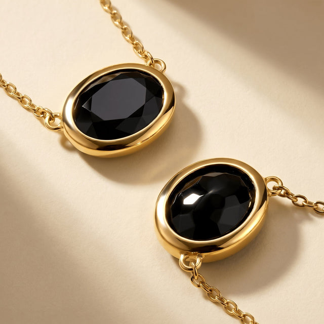 Two yellow-gold-plated black obsidian pendants lie diagonally on a clean pale beige surface.