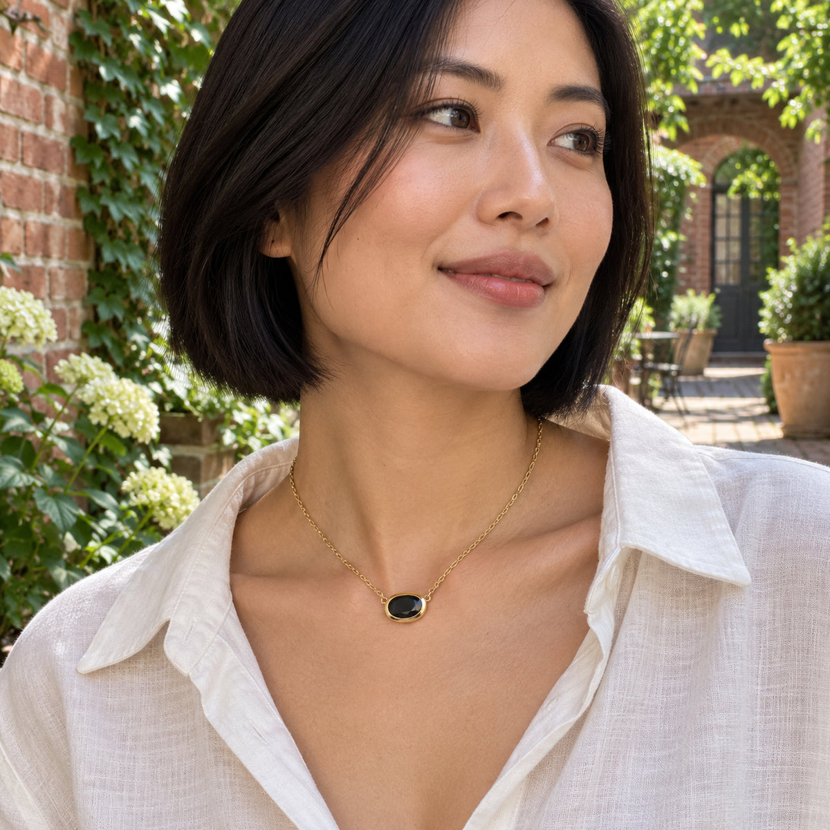 A woman in an ivory linen shirt wears a yellow-gold-plated black obsidian necklace in a leafy brick courtyard.
