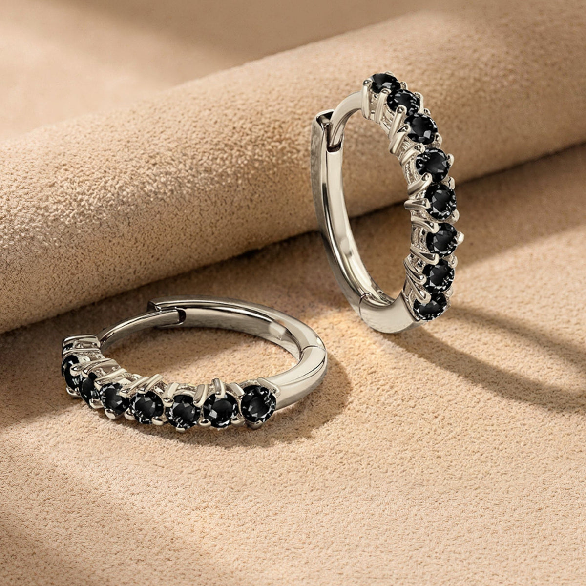White rhodium plated hoop earrings lie on beige suede with their black stones facing forward.