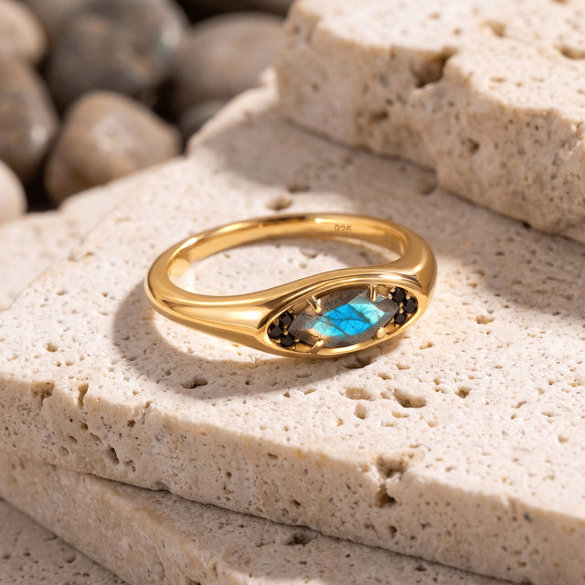 A yellow gold vermeil ring lies on textured pale stone with a bright blue flash across the center gem.