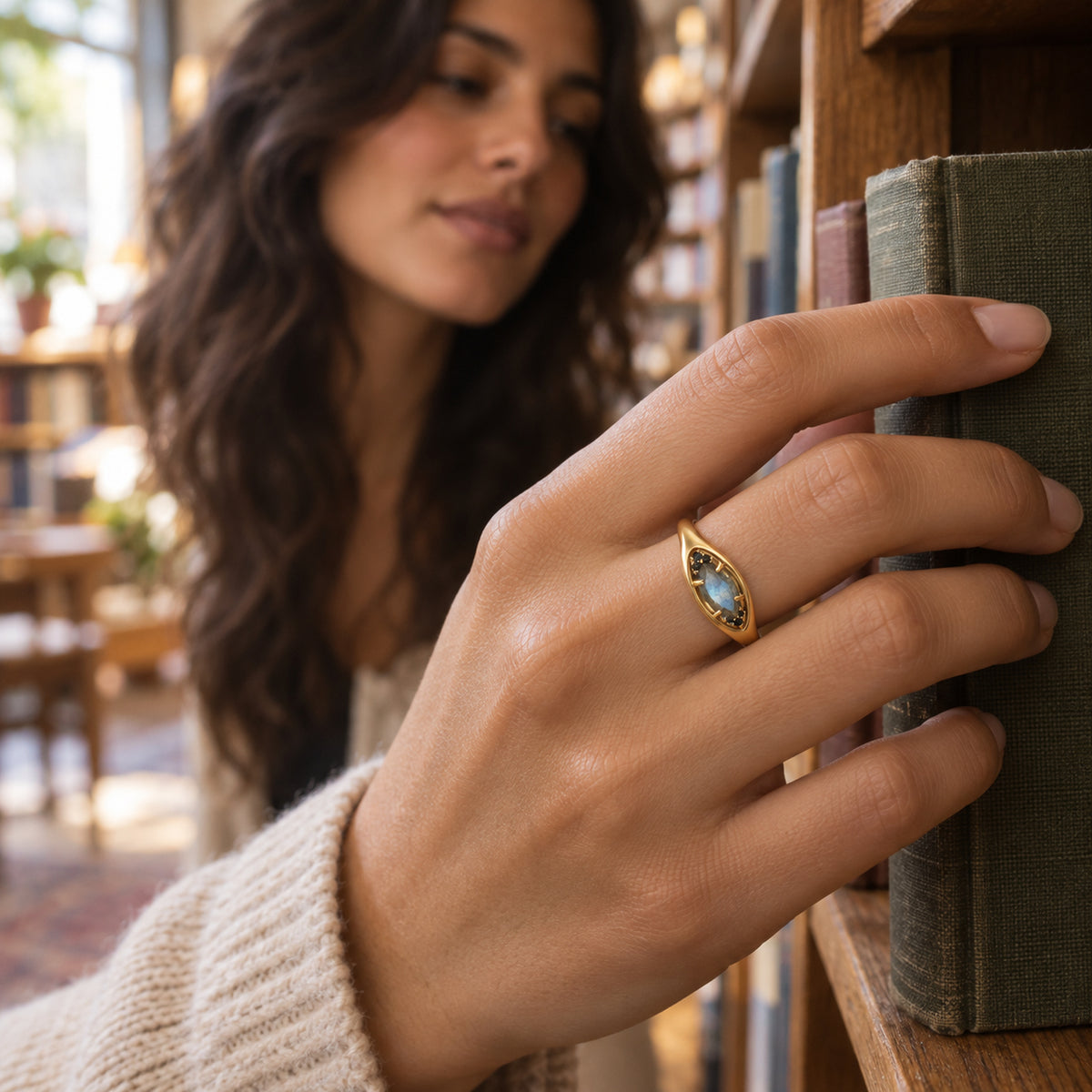 A woman with long dark hair wears a gold-toned labradorite ring as her hand rests against a clothbound book in a bookshop.