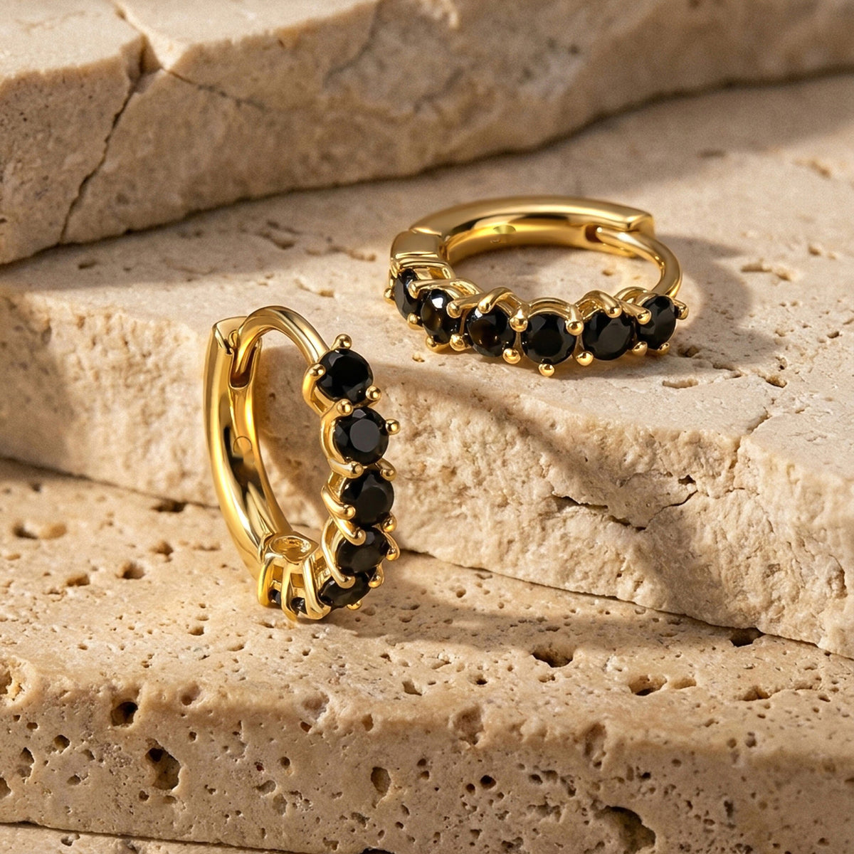 Two gold tone black onyx hoop earrings rest on textured beige stone.