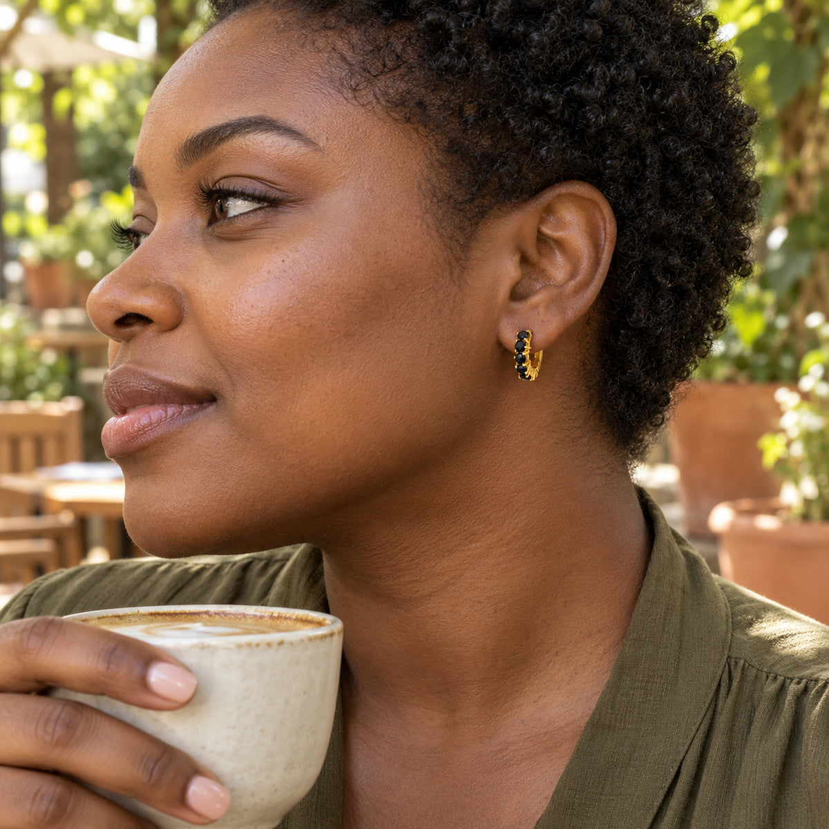 A woman with short curls wears a yellow gold plated black onyx hoop while holding a ceramic cup.
