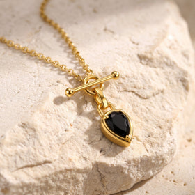 A gold-tone oval-link necklace and black pear-shaped pendant rest across a textured pale stone surface.